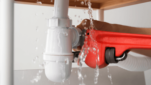 Water leaking from a pipe under a sink while someone tightens it with a red wrench