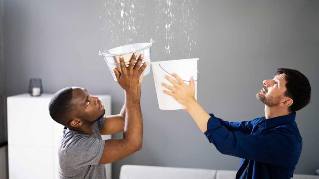 Two men using buckets to catch water from a leaking ceiling.