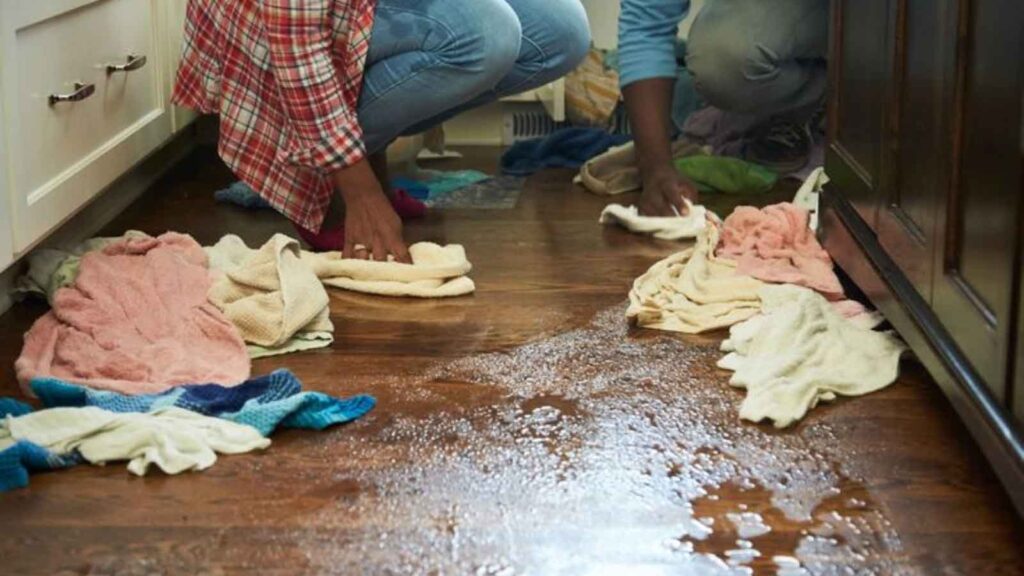 Two people use towels to soak up water from a leaking kitchen floor.
